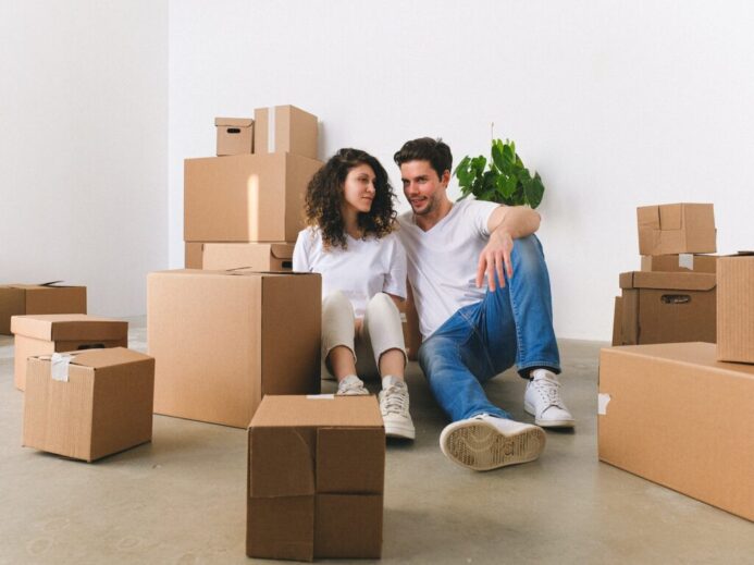 Full body of cheerful couple talking while resting on floor near belongings with carton boxes after moving into new apartment